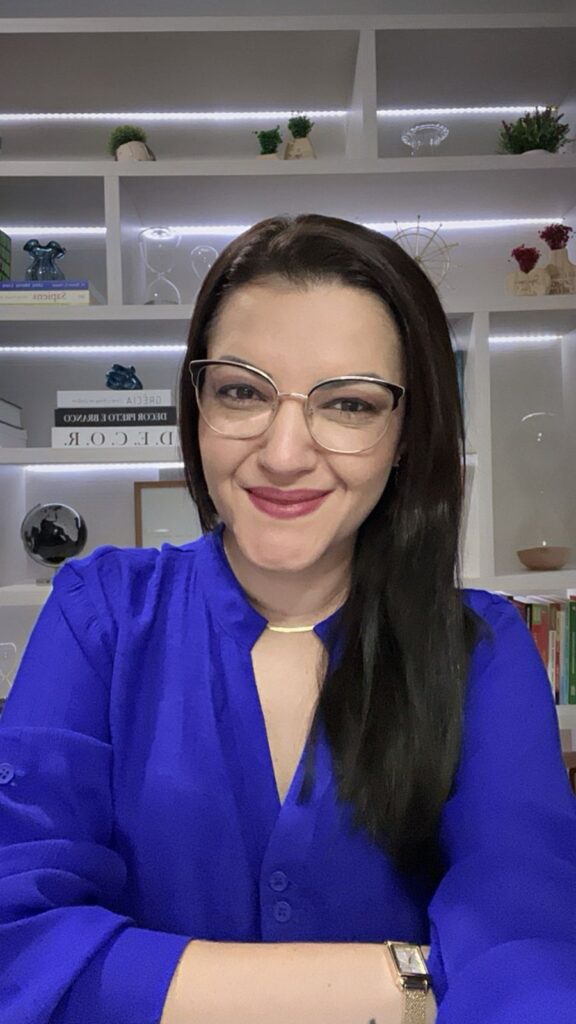 Uma foto minha, estou usando cabelos pretos e longos, com óculos, uma blusa social azul e no meu fundo tem alguns nichos com livros e algumas decorações