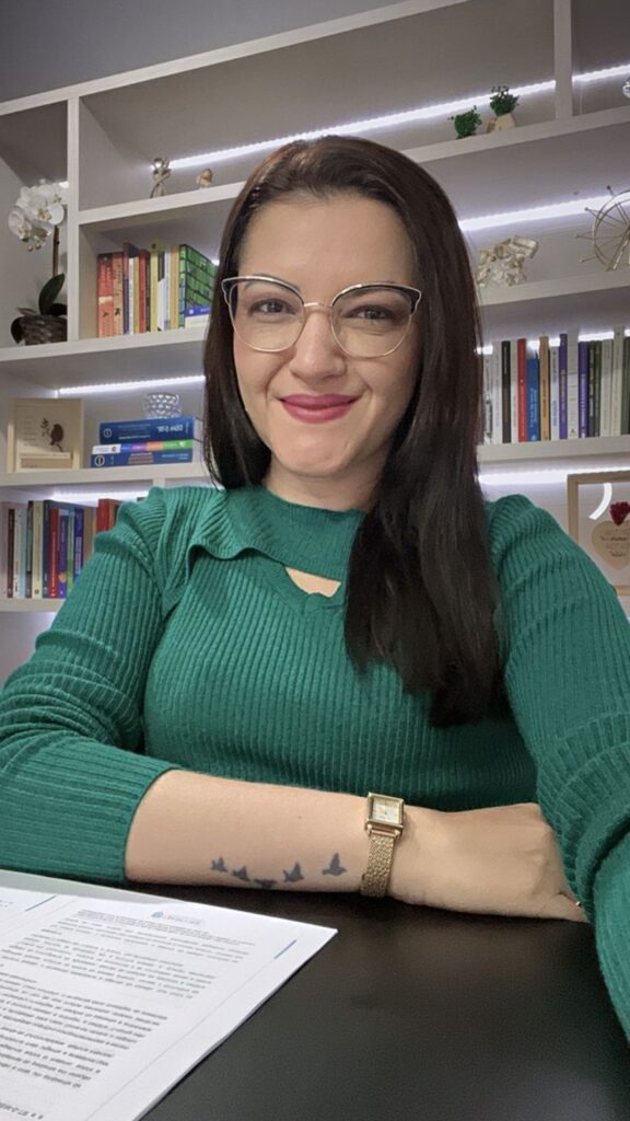 Uma foto minha, estou usando cabelos pretos e longos, com óculos, uma blusa verde e no meu fundo tem alguns nichos com livros e algumas decorações